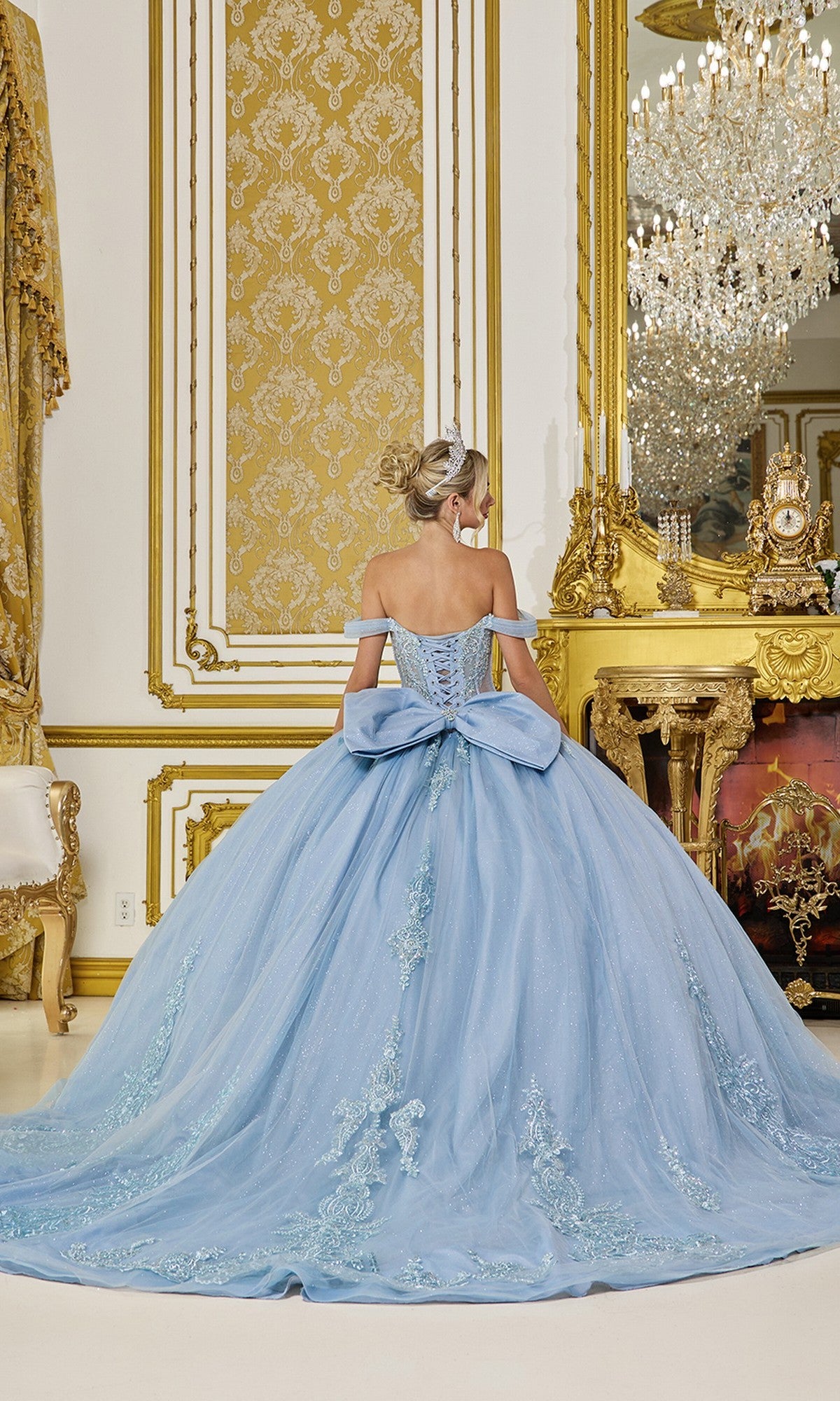 A woman wearing the Quinceanera Dress 5063 By Dancing Queen, featuring a light blue design with a sheer corset bodice and large back bow, stands in an ornate gold and white room, facing away from the camera.